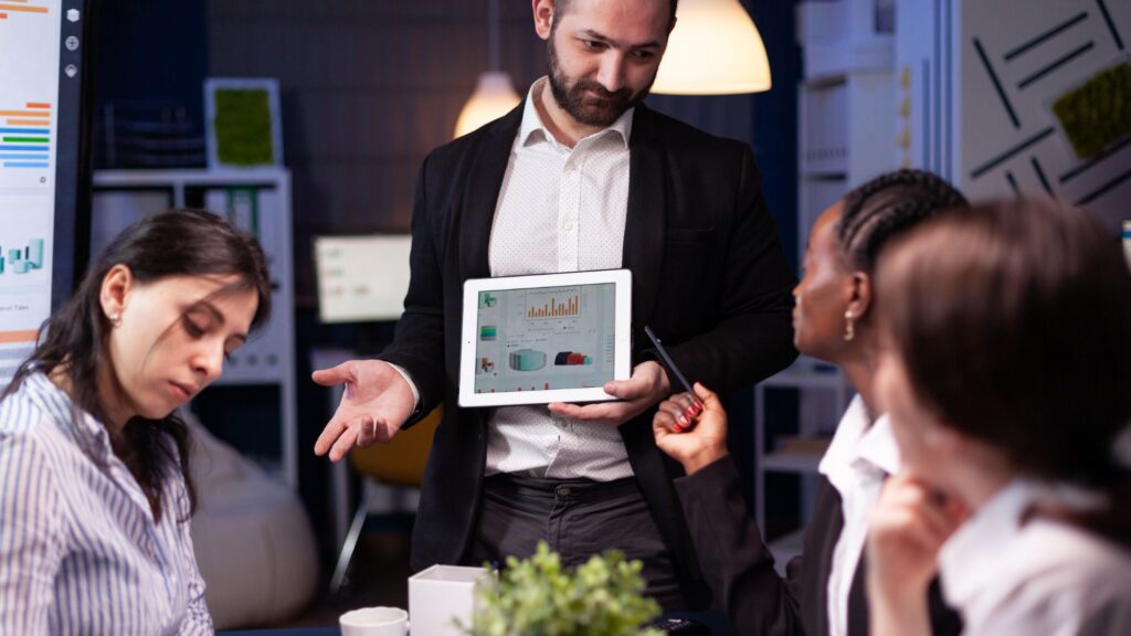  a man holds a tablet as a group of people eagerly gather around, seeking a competitive edge in digital marketing